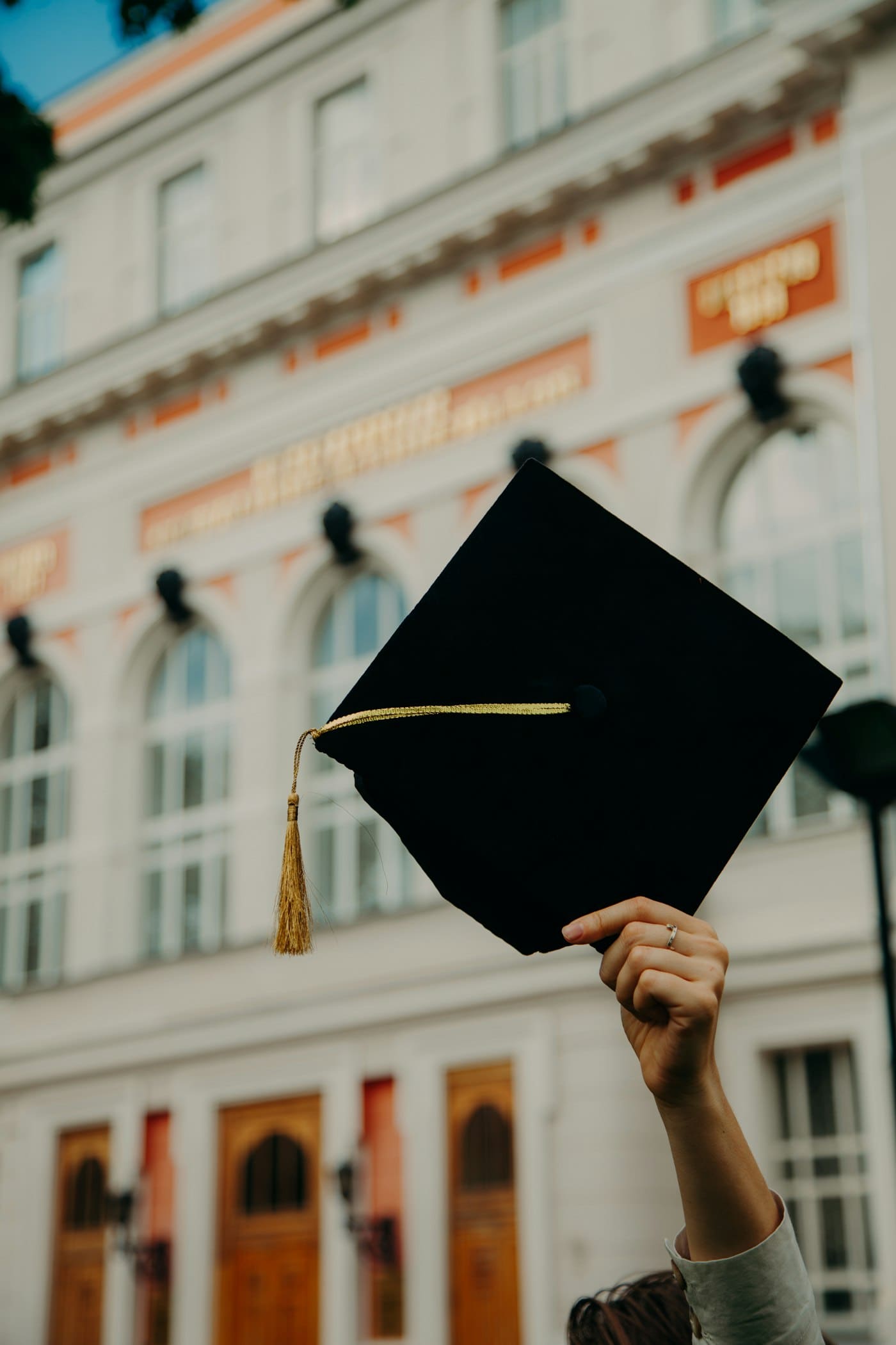 A graduate holds up their cap outside a historic university building